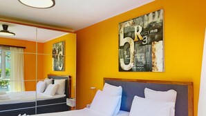 Ett hotellrum med en stor säng, ett sängbord, en spegel och en vägg prydd med ett stort konstverk.