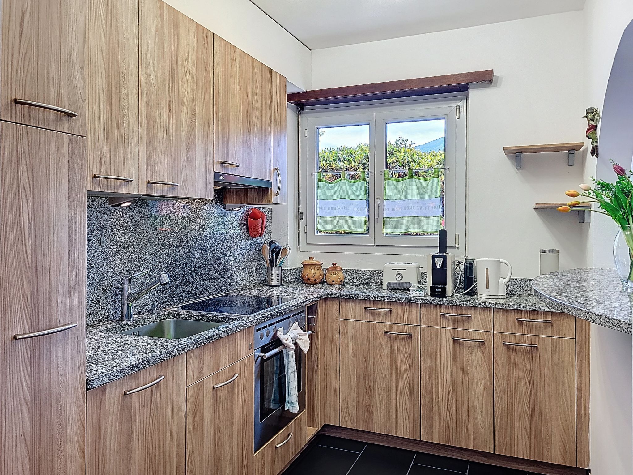 Una cocina con armarios de madera, encimera de granito, horno empotrado, ventana con vistas a zonas verdes y un estante con objetos decorativos.
