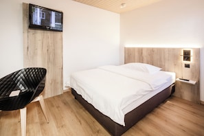 A modern hotel room with a bed, a flat-screen TV mounted on a wooden panel, a bedside table with a lamp, and a black chair.