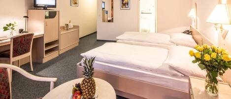 Une chambre d’hôtel avec un lit, un bureau avec un ordinateur, une chaise, une petite table avec des fruits et un vase avec des fleurs.