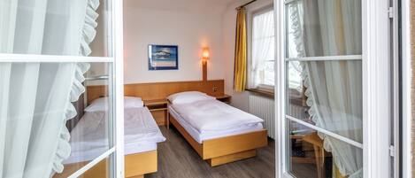 A hotel room with two wooden beds, a nightstand, a wall-mounted lamp, a window with curtains, and a framed picture on the wall.