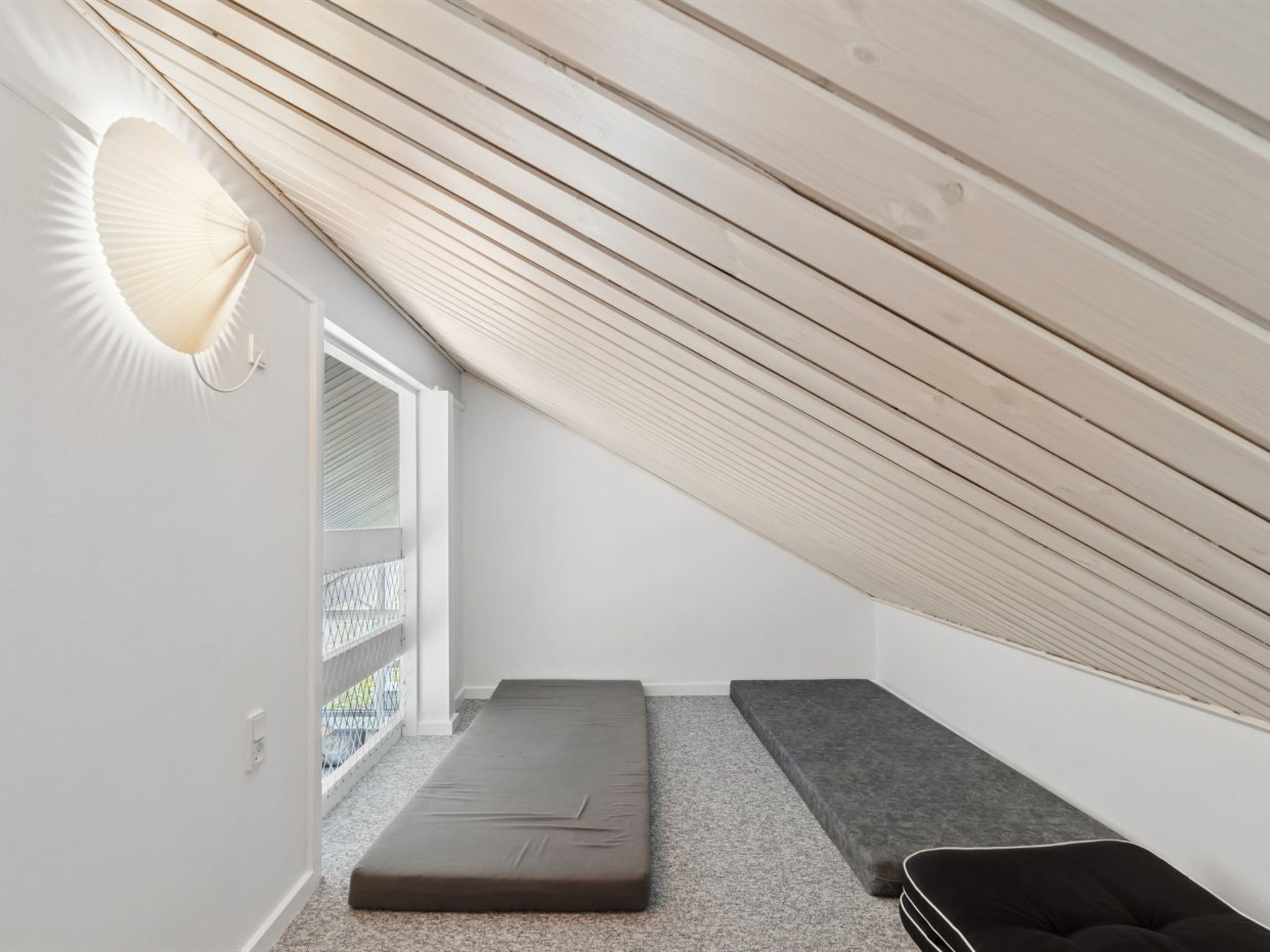 Ein kleiner Raum mit geneigter Decke, einem Fenster, einer Bank und einer Wandleuchte.