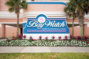 A sign for Bay Watch Resort & Conference Center with a clear view of the building's exterior and a flower bed in front. - Cozy 2-bedroom condo on the 10th Floor, located in fantastic North Myrtle Beach (North Myrtle Beach)
