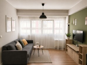 Ein modernes Wohnzimmer mit einem grauen Sofa, einem runden Couchtisch, einem Fernseher und einem großen Fenster mit transparenten Vorhängen.