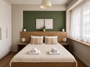 A bedroom with a wooden headboard, two bedside tables with lamps, a bed with a striped blanket and rolled towels, a green accent wall, and two framed pictures. - Comfortable 1BR Apartment 5 min to Ladadika (Thessaloniki)