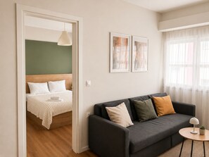 A modern living room with a sofa, a small side table with a lamp, and a bed with a wooden headboard. - Comfortable 1BR Apartment 5 min to Ladadika (Thessaloniki)
