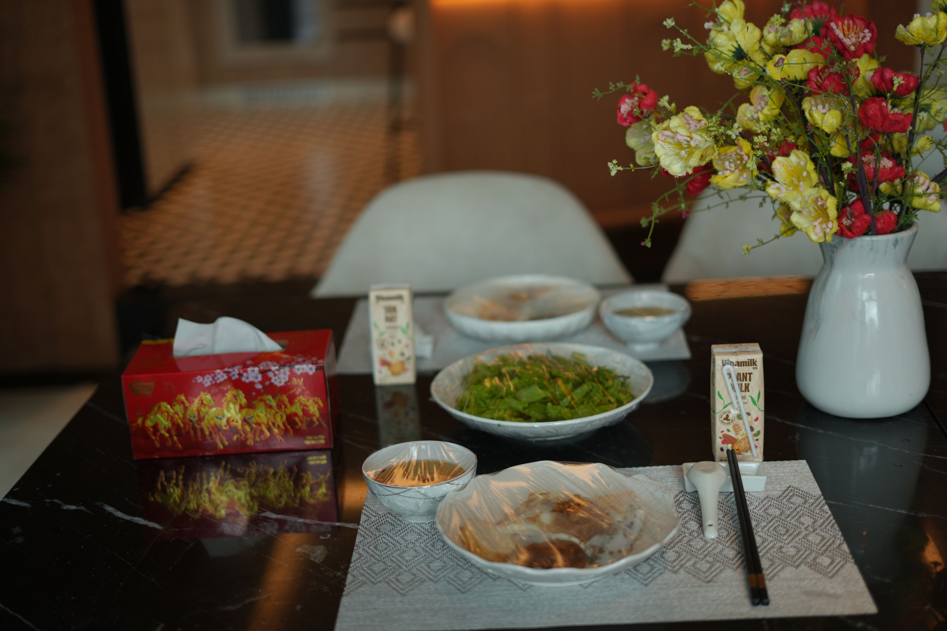 A table with a vase of flowers, a box of tissues, and various dishes.