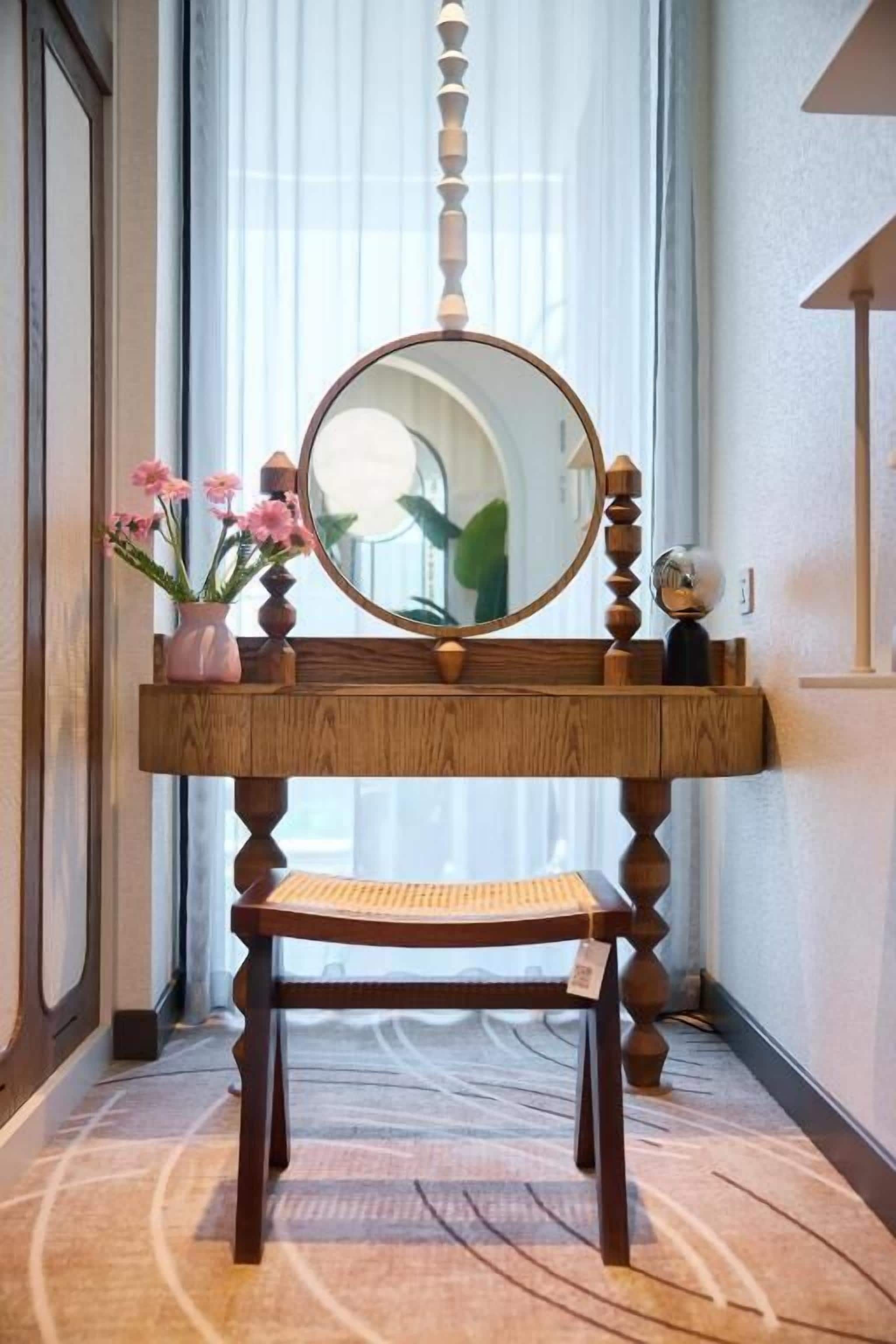 A wooden vanity with a mirror, a small stool, and a vase with pink flowers.