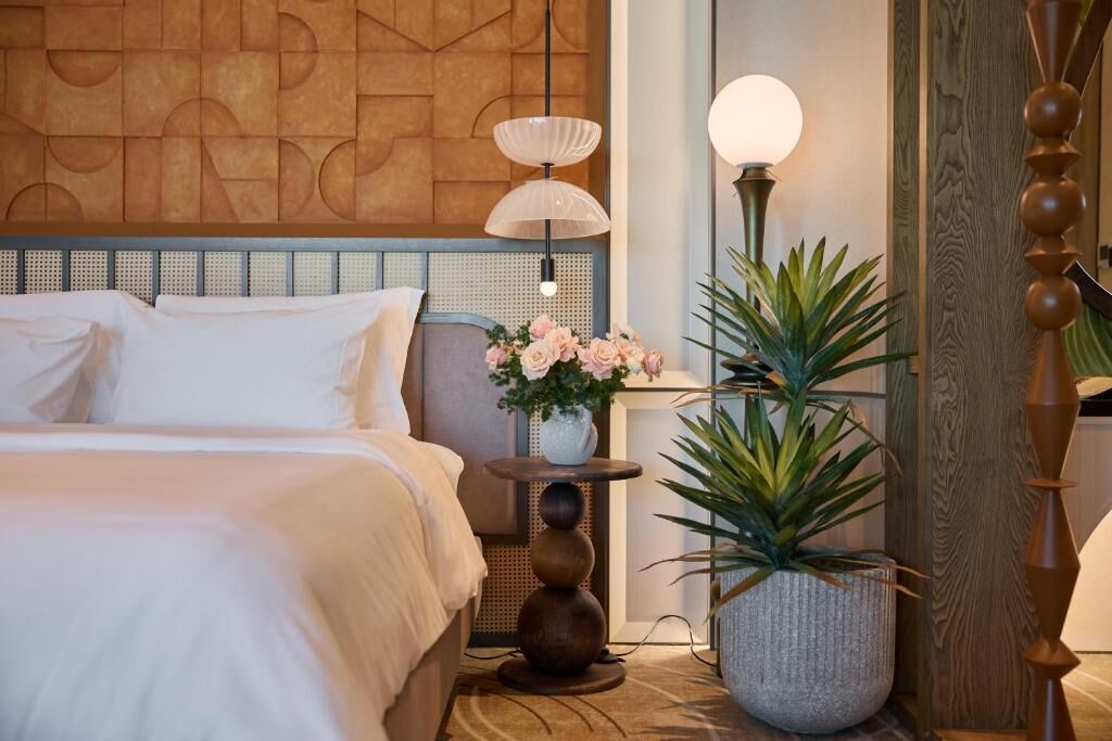 A neatly made bed with white linens, a wall with a geometric pattern, a round table with a vase of flowers, and a tall floor lamp.