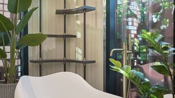 A modern bathroom with a white bathtub, a glass shelf unit, and potted plants.