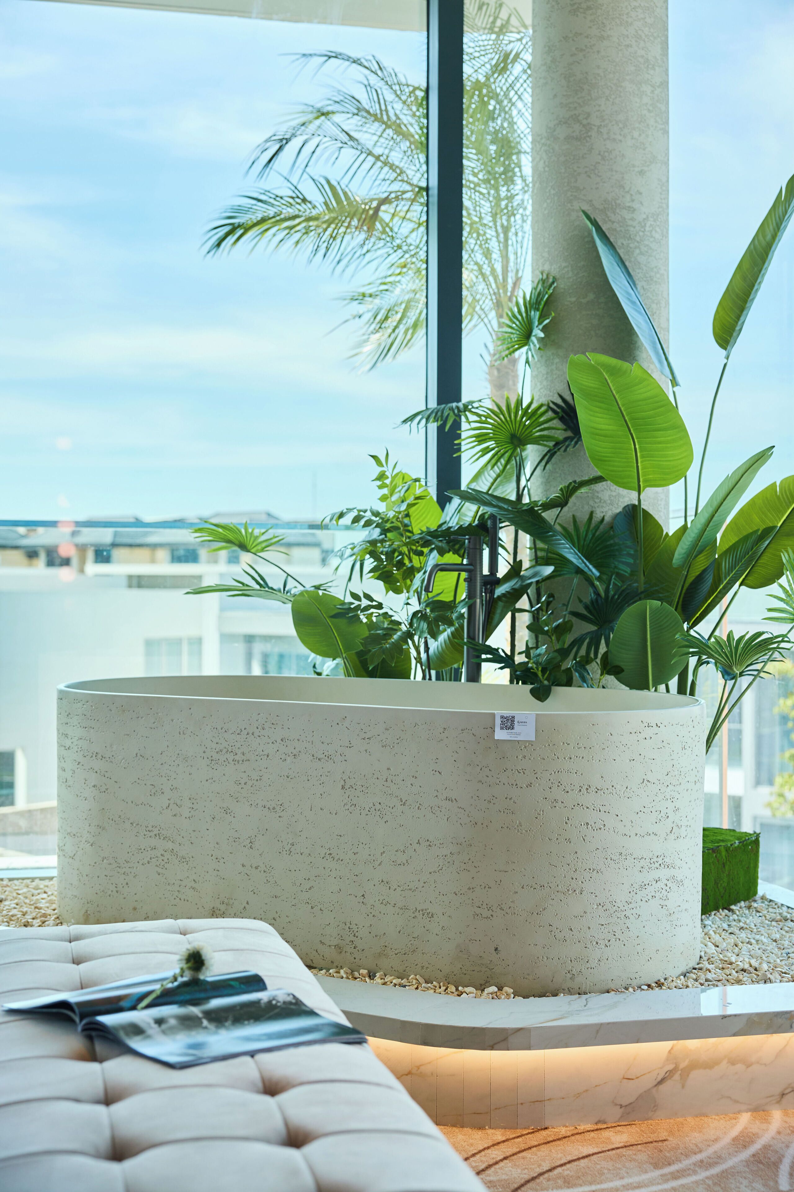 Un espace intérieur moderne, doté d’un grand bac en béton rempli de différentes plantes vertes, d’un banc blanc rembourré et offrant un aperçu d’un paysage urbain à travers de grandes fenêtres.