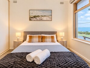 A hotel room with a bed, two bedside lamps, a view of the beach, and a painting on the wall.