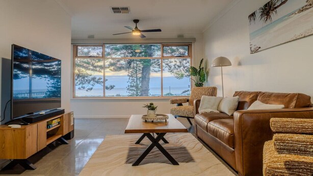 A living room with a large window, a ceiling fan, a brown leather sofa, a wooden coffee table, and a TV on a wooden stand.