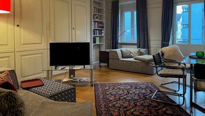 Moderni olohuone, jossa on litteänäyttöinen televisio, sohva, nojatuoli, lasinen sohvapöytä, kirjahylly ja suuri ikkuna, jossa on verhot.