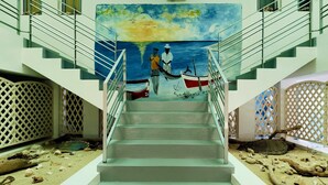 Una escalera con un mural de dos figuras y barcos, rodeada de un suelo arenoso con conchas dispersas.