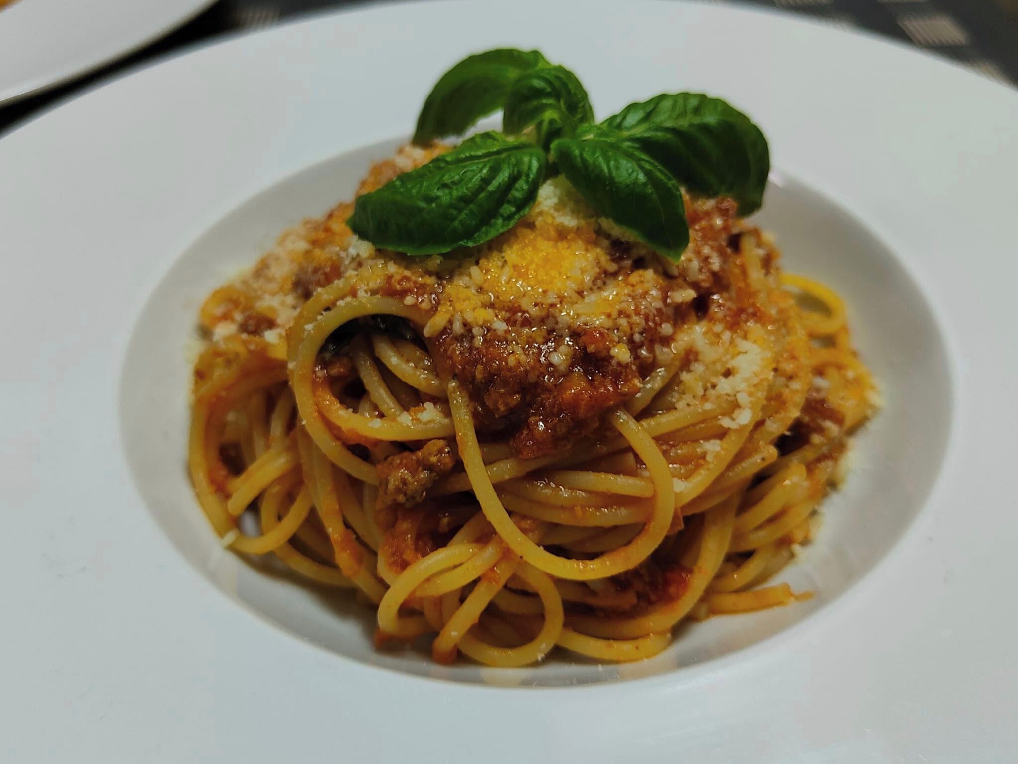 Een bord spaghetti met vleessaus en geraspte kaas, versierd met een basilicumblaadje.