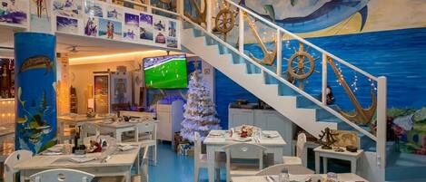 Un restaurante de temática náutica con una escalera, mesas, sillas, un árbol de Navidad y un mural de un barco a vela.
