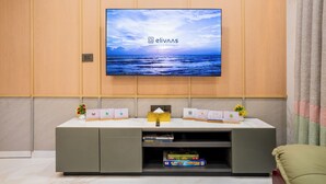 A modern living room with a flat-screen TV mounted on a wooden wall, a grey entertainment unit below, and a view of the sea on the screen. - Alaya Stays Rosalia 3BHK Villa with Pool (Lonavala)