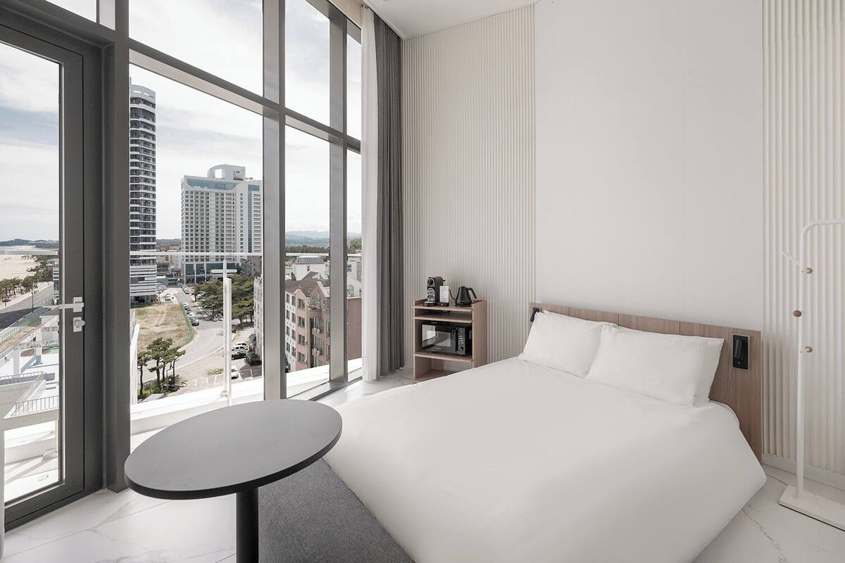 Ett modernt hotellrum med en stor säng, ett litet runt bord, utsikt över staden genom stora fönster och en inbyggd hylla med en vattenkokare och en telefon.
