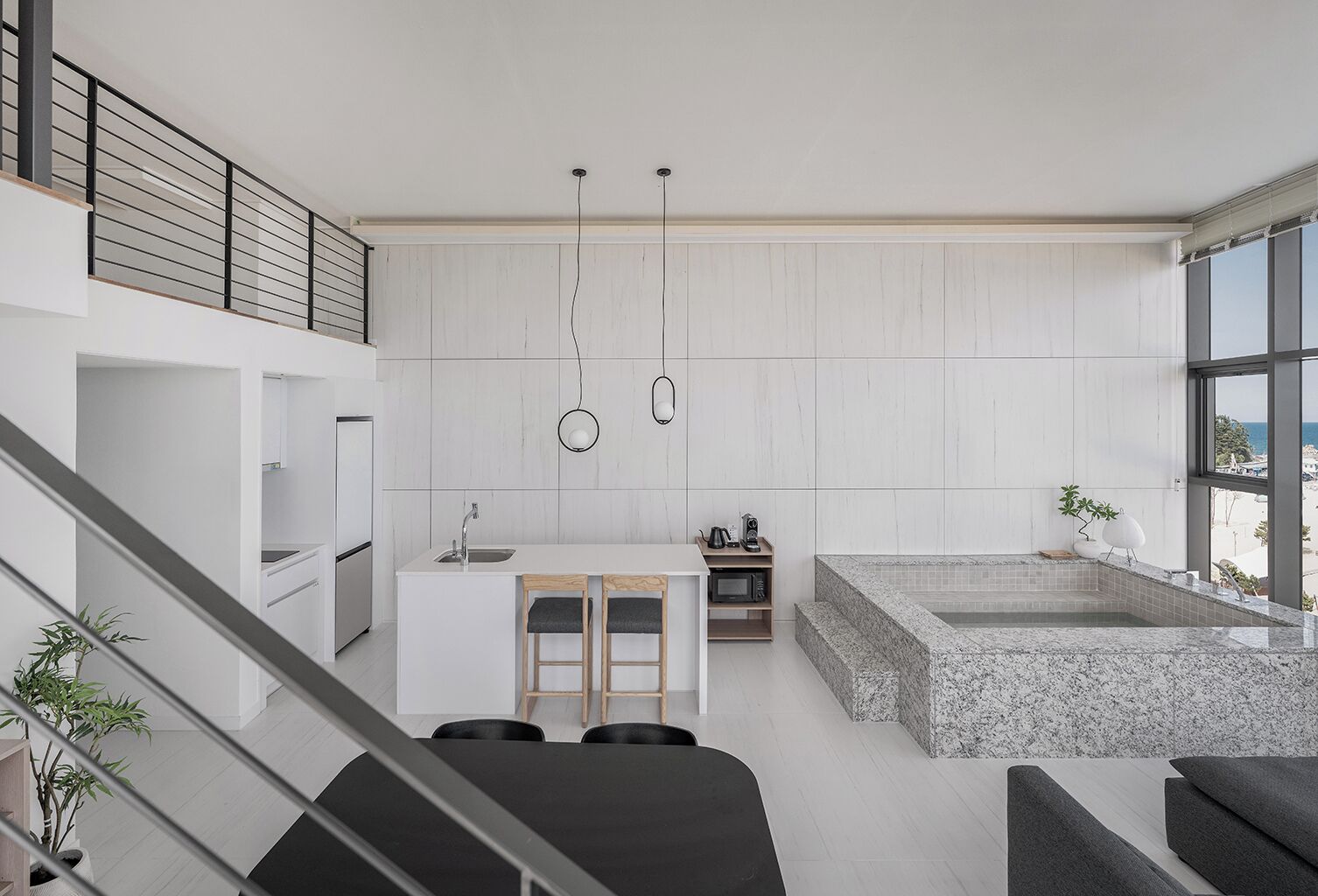 A modern kitchen with a central island, a dining area with chairs, a large window with a view, and a staircase leading to an upper level.