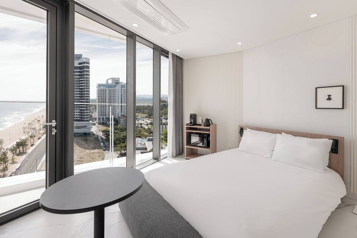 Ett modernt hotellrum med en stor säng, ett litet bord, utsikt över stranden och staden samt en minibar.