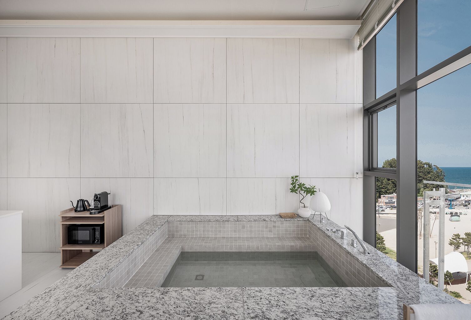 Un baño moderno con una bañera grande, un estante de madera con una tetera y vista a la playa a través de amplias ventanas.