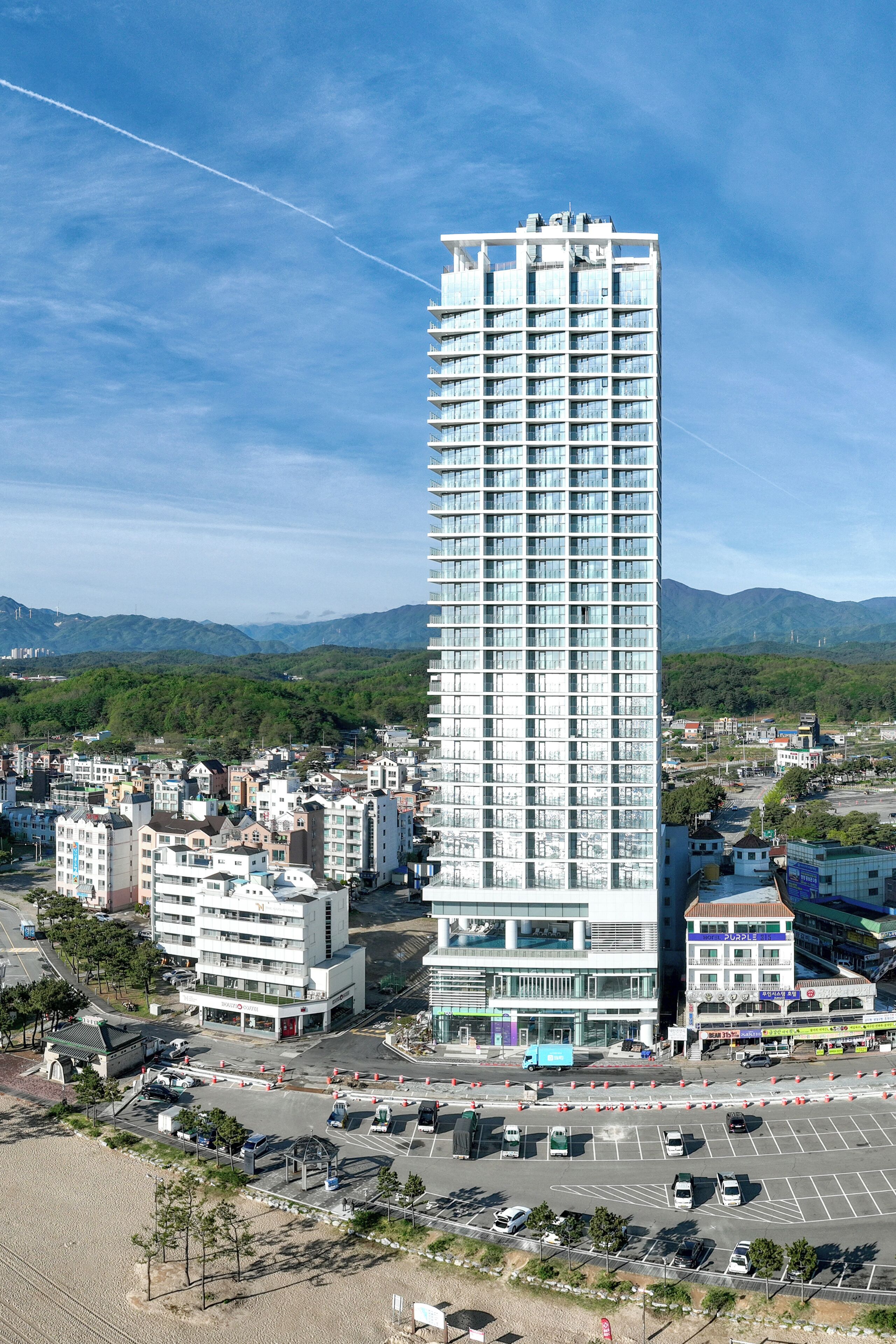 A tall, modern building with multiple floors, surrounded by other buildings and a parking lot.