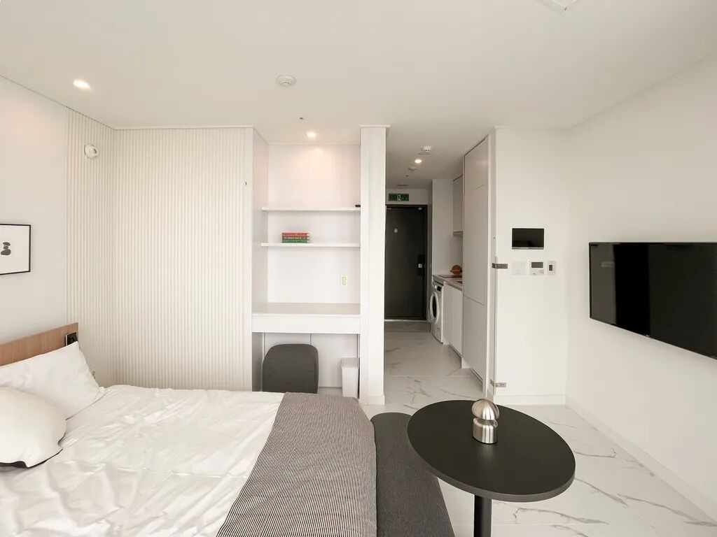 A modern hotel room with a bed, a small table, a TV, and a kitchenette in the background.