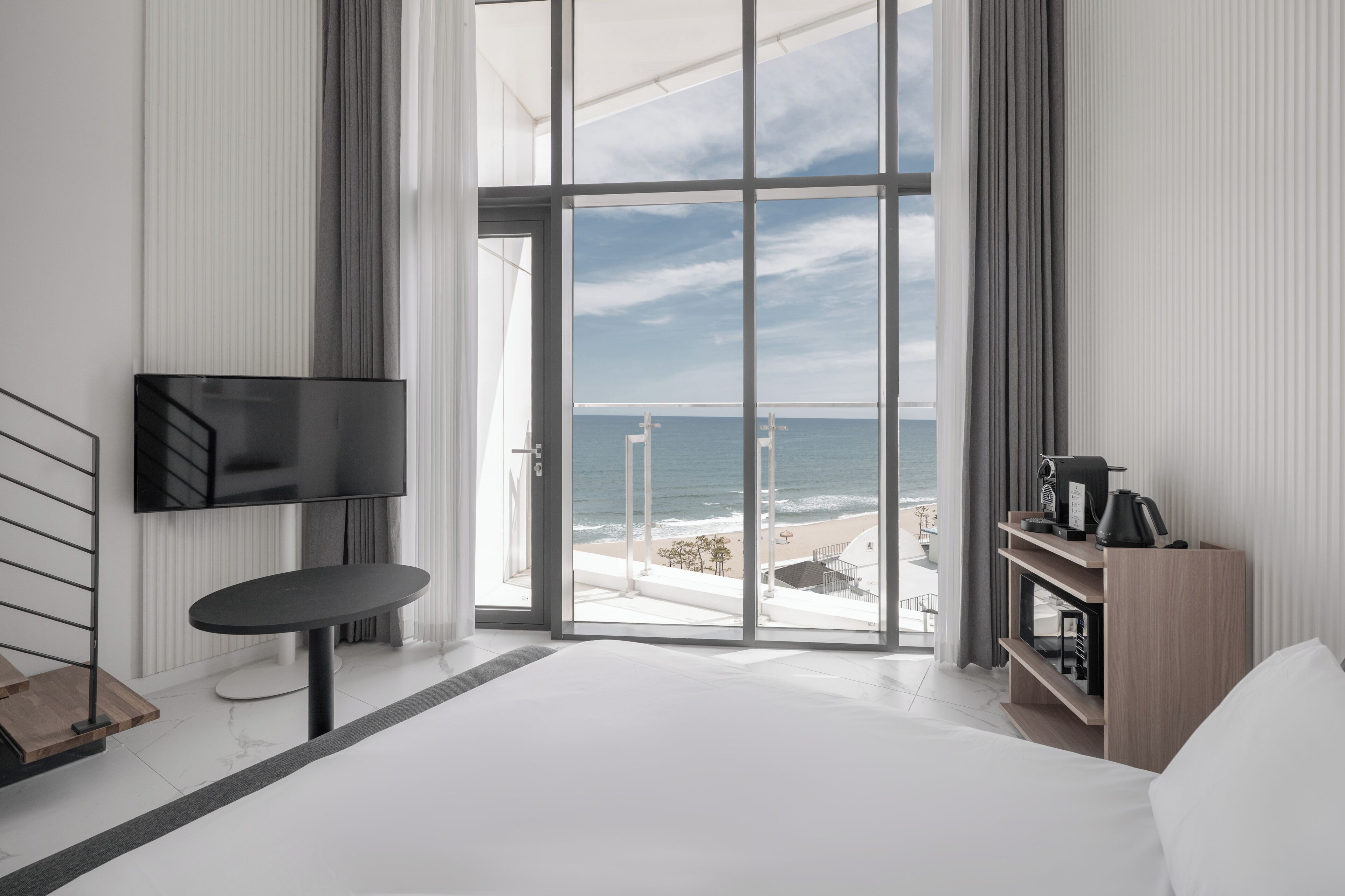 A modern hotel room with a large window, a flat-screen TV, a small table, a bed, and a wooden shelf.