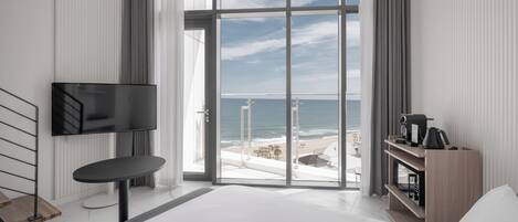 A modern hotel room with a large window, a flat-screen TV, a small table, a bed, and a wooden shelf.