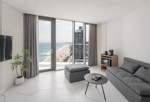 A modern living room with a large window, a grey sofa, a small table, and a bookshelf. - Plott The Gak Yangyang (Yangyang)
