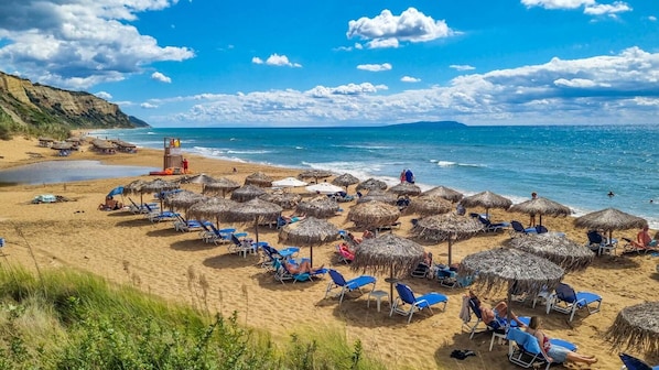 Ein Strand mit reetgedeckten Sonnenschirmen und blauen Liegestühlen, einem Rettungsturm und freiem Blick auf das Meer.
