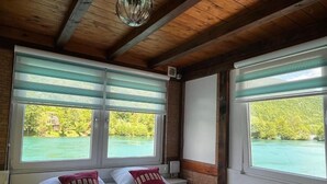 Quarto com duas camas, janelas amplas com persianas, teto de madeira e vista para uma paisagem verde.