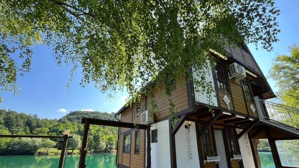 Uma casa de madeira com varanda, uma parede de pedra e uma mesa com cadeiras em um cenário à beira do lago.
