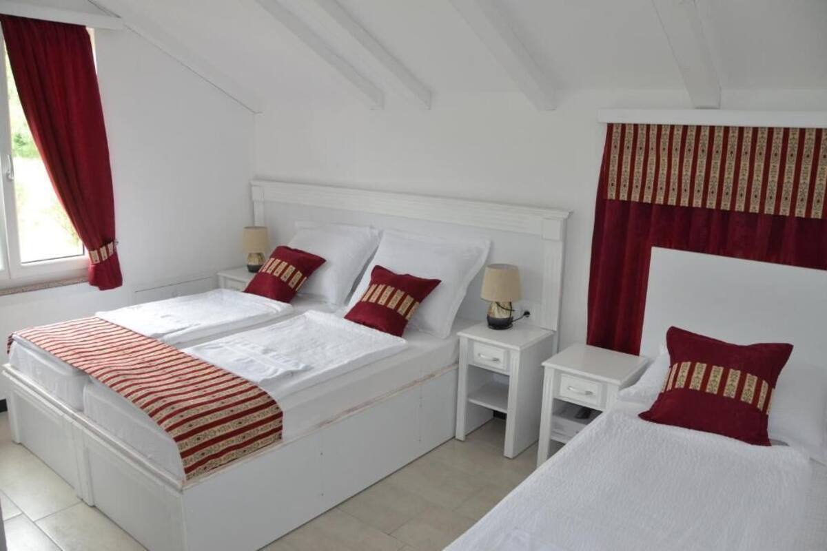 Una cama doble con dos almohadas, una mesita de noche con lámpara y una ventana con cortinas rojas.