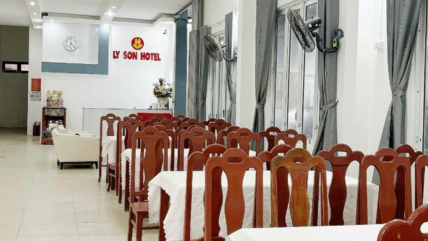 Ένας χώρος εστίασης σε ένα ξενοδοχείο, με σειρές από ξύλινα καθίσματα και τραπέζια, ένα ρολόι και μια πινακίδα με την επιγραφή «LY SON HOTEL».