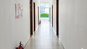 Ένας μακρύς, στενός διάδρομος με λευκούς τοίχους και πλακάκι στο δάπεδο, που οδηγεί σε ένα φωτεινό δωμάτιο με θέα στο πράσινο του εξωτερικού χώρου.