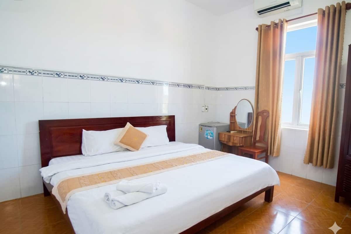 A hotel room with a wooden bed, white bedding, a small dresser with a mirror, and a window with curtains.