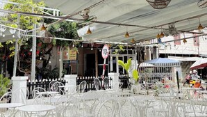 Ένα υπαίθριο καφέ με λευκές μεταλλικές καρέκλες και τραπέζια, κρεμαστά φωτιστικά και μια σκεπαστή περιοχή με μια πινακίδα που υποδεικνύει περιορισμό ύψους.