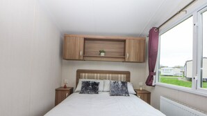 A bedroom with a bed, wooden headboard, bedside tables, a window with curtains, and a view of the outside. - 49 Cader Idris, Hafan-y-mor Holiday Park (Pwllheli)