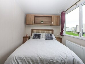 A bedroom with a bed, wooden headboard, bedside tables, a window with curtains, and a view of the outside. - 49 Cader Idris, Hafan-y-mor Holiday Park (Pwllheli)