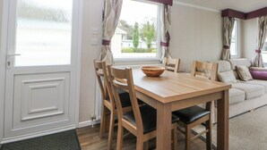 A dining area with a wooden table and chairs, a window with curtains, and a sofa in the background. - 49 Cader Idris, Hafan-y-mor Holiday Park (Pwllheli)