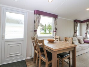 A dining area with a wooden table and chairs, a window with curtains, and a sofa in the background. - 49 Cader Idris, Hafan-y-mor Holiday Park (Pwllheli)