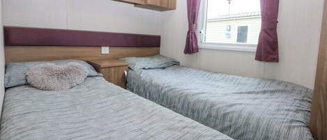 Une chambre avec deux lits, des armoires en bois au-dessus, une fenêtre avec des rideaux et une prise électrique visible sur le mur.