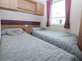 A bedroom with two beds, wooden cabinets above, a window with curtains, and a visible power outlet on the wall.