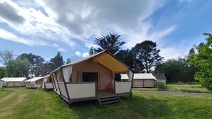 A series of camping tents with a flat roof and a small porch area, set in a grassy field with trees in the background.