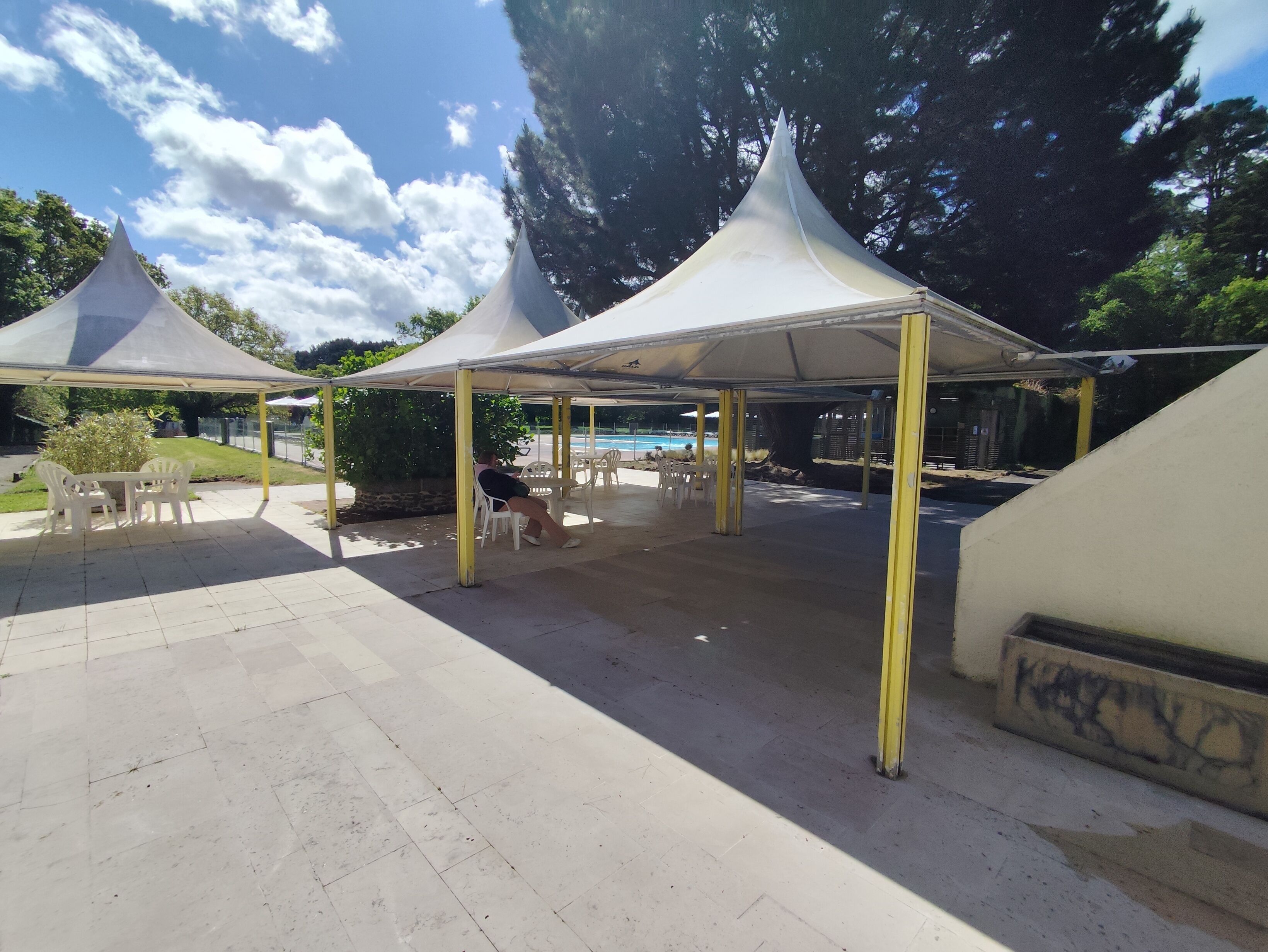 Ein überdachter Außenbereich mit Tischen und Stühlen, im Hintergrund ein Pool und Bäume rund um den Platz.