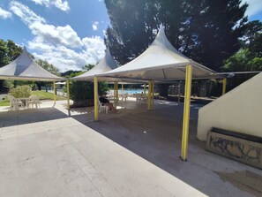 Un espace extérieur abrité, doté de tables et de chaises, avec une piscine en arrière-plan et des arbres qui entourent l’ensemble.