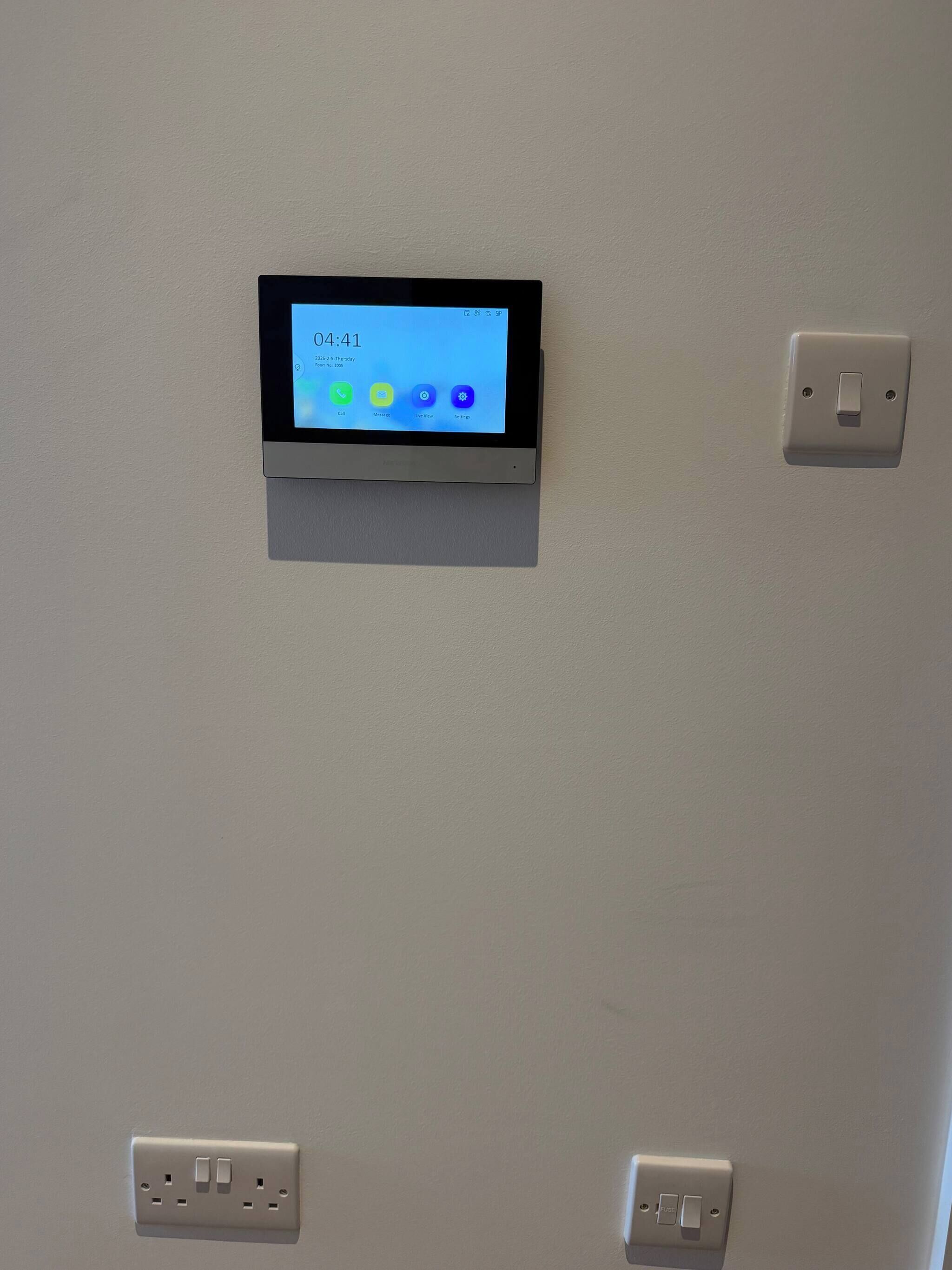 Un reloj digital con pantalla que muestra la hora, la fecha y la información meteorológica, montado en una pared con dos tomas de corriente debajo.