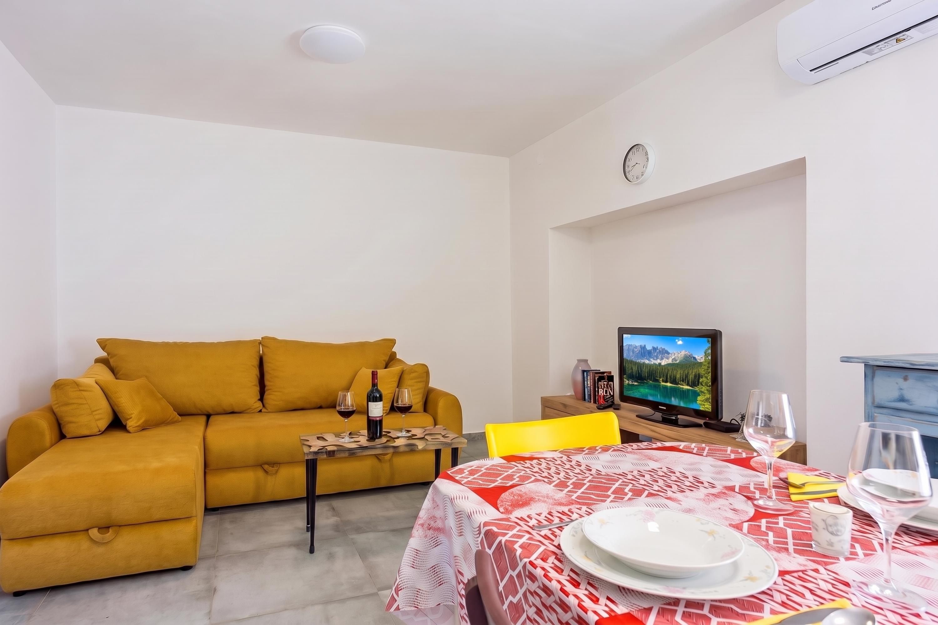 A living room with a sofa, a dining table set with plates and glasses, a television, and a clock on the wall.